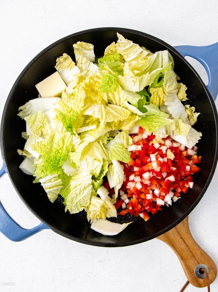 A top-down view of a black and blue skillet containing chopped napa cabbage, diced red bell pepper, and diced white onion on a light surface.