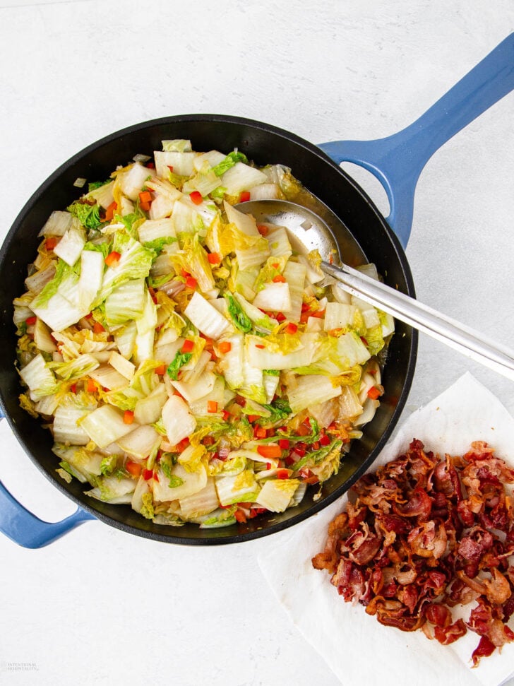 A skillet filled with sautéed cabbage and diced red peppers sits on a white surface beside a pile of cooked, crispy bacon on a paper towel. Metal tongs rest on the pan’s edge.