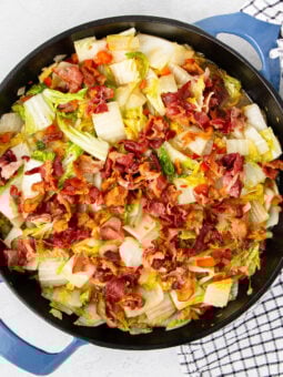 A skillet filled with cooked cabbage, red peppers, and crispy bacon pieces, placed on a white surface next to a black-and-white checkered towel.