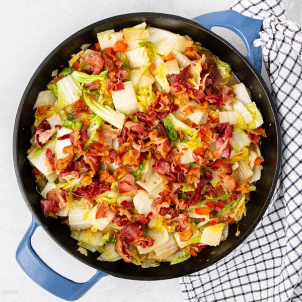 A skillet filled with cooked cabbage, red peppers, and crispy bacon pieces, placed on a white surface next to a black-and-white checkered towel.