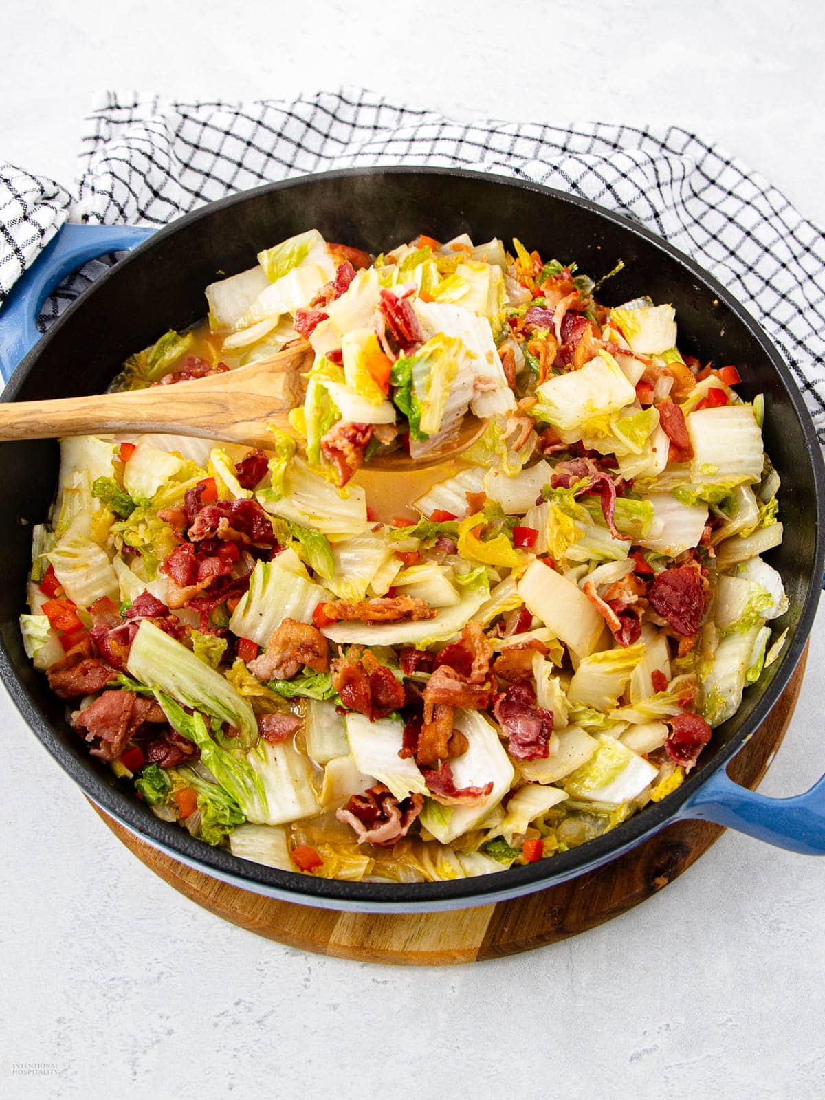 A skillet filled with cooked chopped cabbage, crispy bacon pieces, and diced red peppers, with a wooden spoon resting inside. The skillet sits on a round wooden board atop a white surface, next to a checkered cloth.