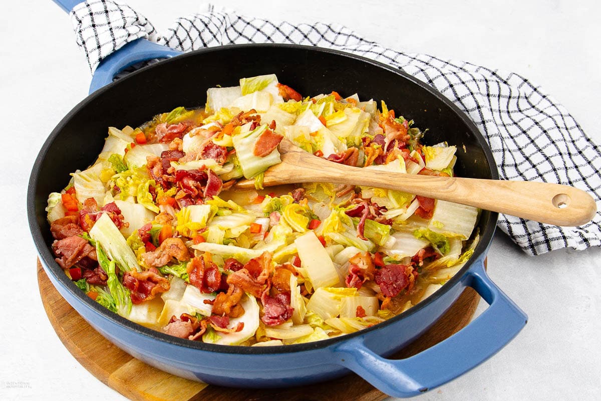 A blue skillet filled with cooked cabbage, carrots, and crispy bacon sits on a wooden board. A wooden spoon rests in the pan, and a black-and-white checkered towel is beside it on a light surface.