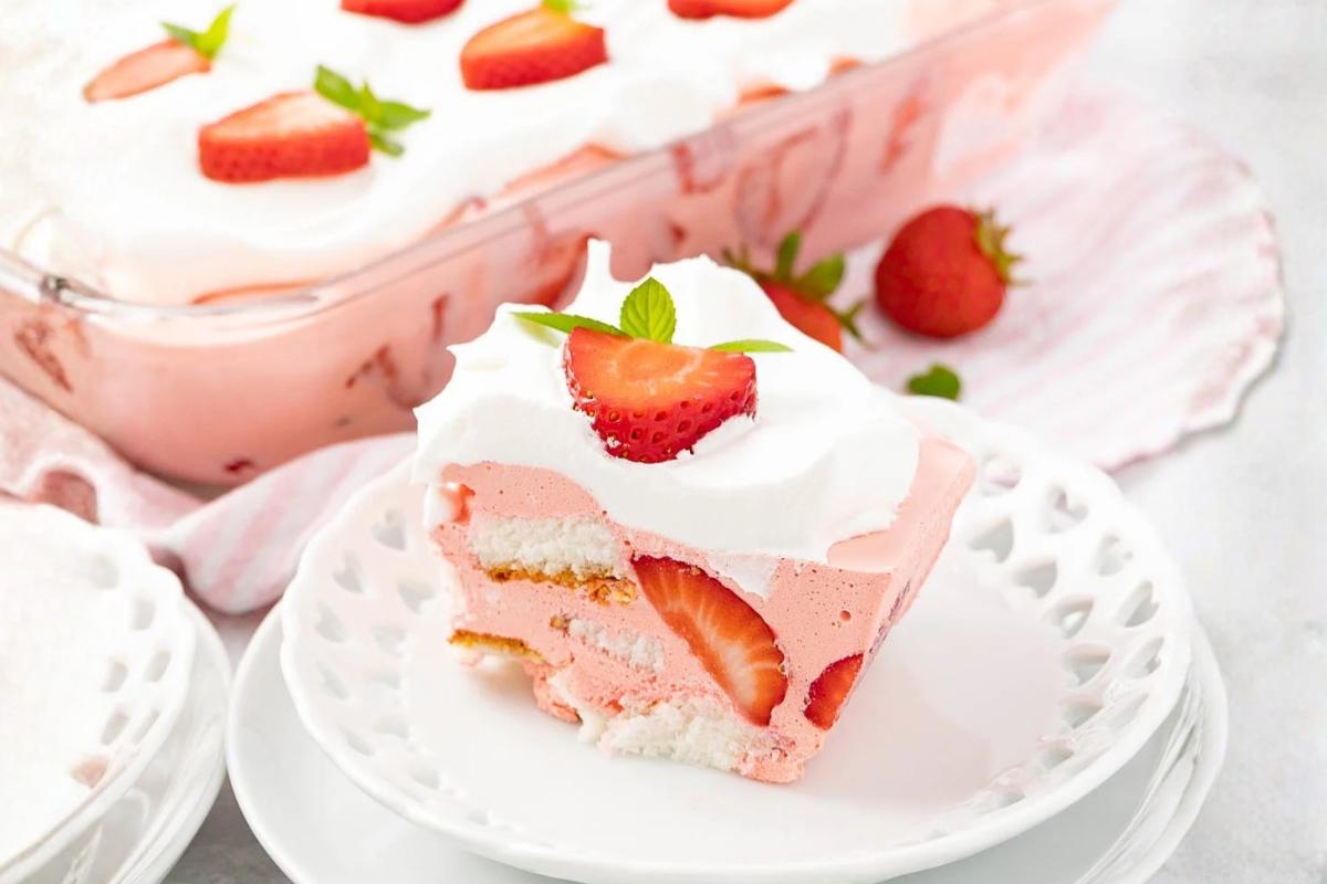 A slice of strawberry dessert with layers of pink mousse, whipped cream, fresh strawberries, and ladyfinger biscuits sits on a white plate. A glass dish with more dessert and whole strawberries are in the background.