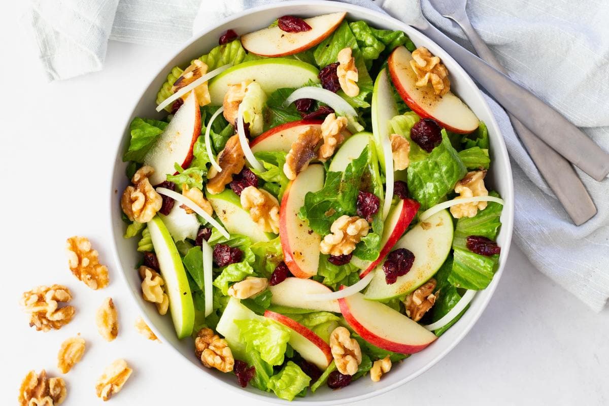 A bowl of fresh salad with romaine lettuce, sliced red and green apples, walnuts, dried cranberries, and thin onion slices, with extra walnuts scattered on the white surface nearby. A fork and knife rest beside the bowl.