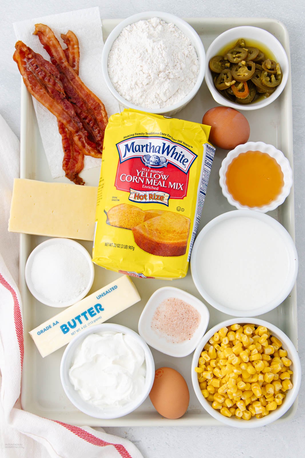 A tray holds ingredients including bacon strips, a block of cheese, flour, jalapeno slices, eggs, cornmeal mix, honey, butter, sour cream, salt, corn kernels, and sugar in separate bowls or containers.