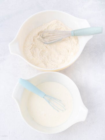 Two white mixing bowls on a light surface. The top bowl contains flour mixture and a whisk; the bottom bowl contains a liquid mixture and a whisk. Both whisks have light blue handles.