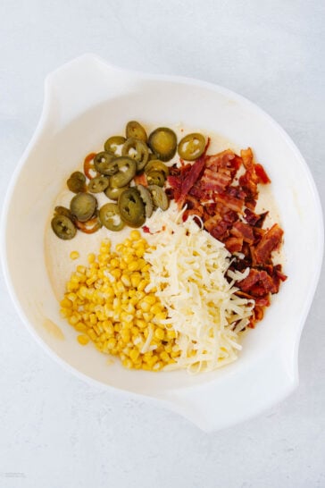 A white mixing bowl containing sliced jalapeños, crumbled bacon, shredded cheese, and corn, arranged in separate sections on a light background.