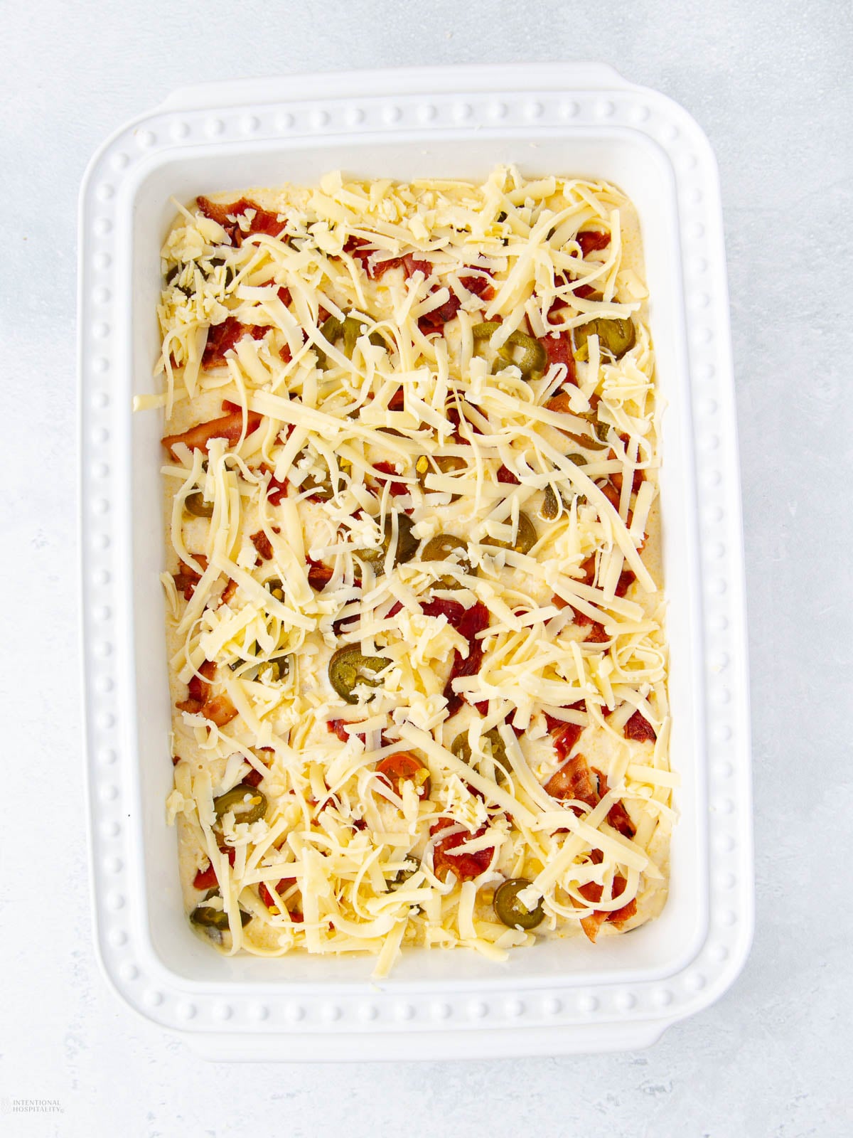 A white baking dish filled with a layered casserole topped with shredded cheese, sliced jalapeños, and diced tomatoes, ready to be baked.
