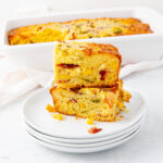 Two slices of savory cornbread with visible pieces of corn, red peppers, and green peppers are stacked on a white plate, with a larger cornbread loaf in a white baking dish in the background.