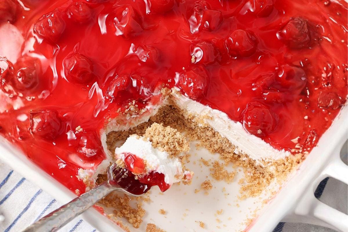 A baking dish with a cherry cheesecake dessert, showing layers of cherry topping, creamy filling, and crumbly crust. A serving has been scooped out with a spoon resting in the dish.
