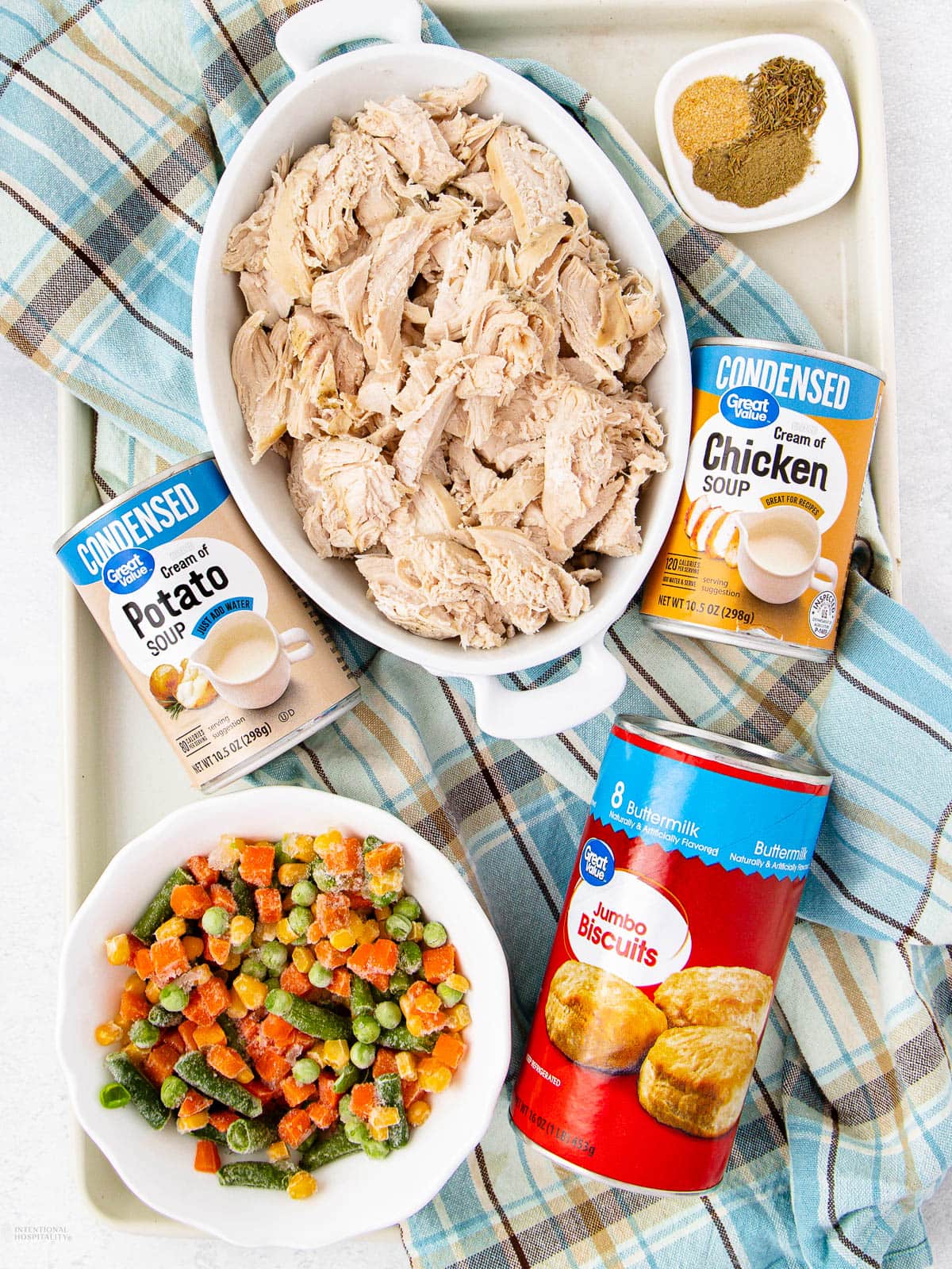 A tray with a dish of shredded chicken, cans of condensed potato and chicken soup, a can of jumbo biscuits, a bowl of mixed vegetables, and a small bowl of spices, all on a blue plaid cloth.