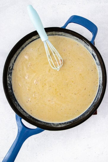 A blue-handled pan filled with creamy, light-colored sauce being whisked with a blue-handled whisk, shown from above on a light surface.