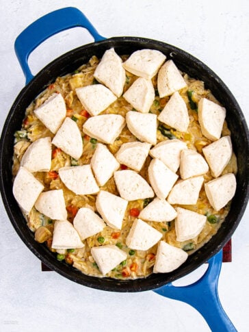 A blue skillet filled with creamy chicken and vegetable mixture, topped with uncooked biscuit dough pieces, ready to be baked.