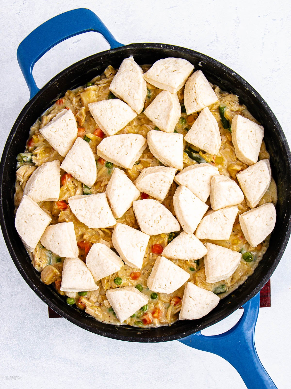 A blue skillet filled with creamy chicken and vegetable mixture, topped with uncooked biscuit dough pieces, ready to be baked.