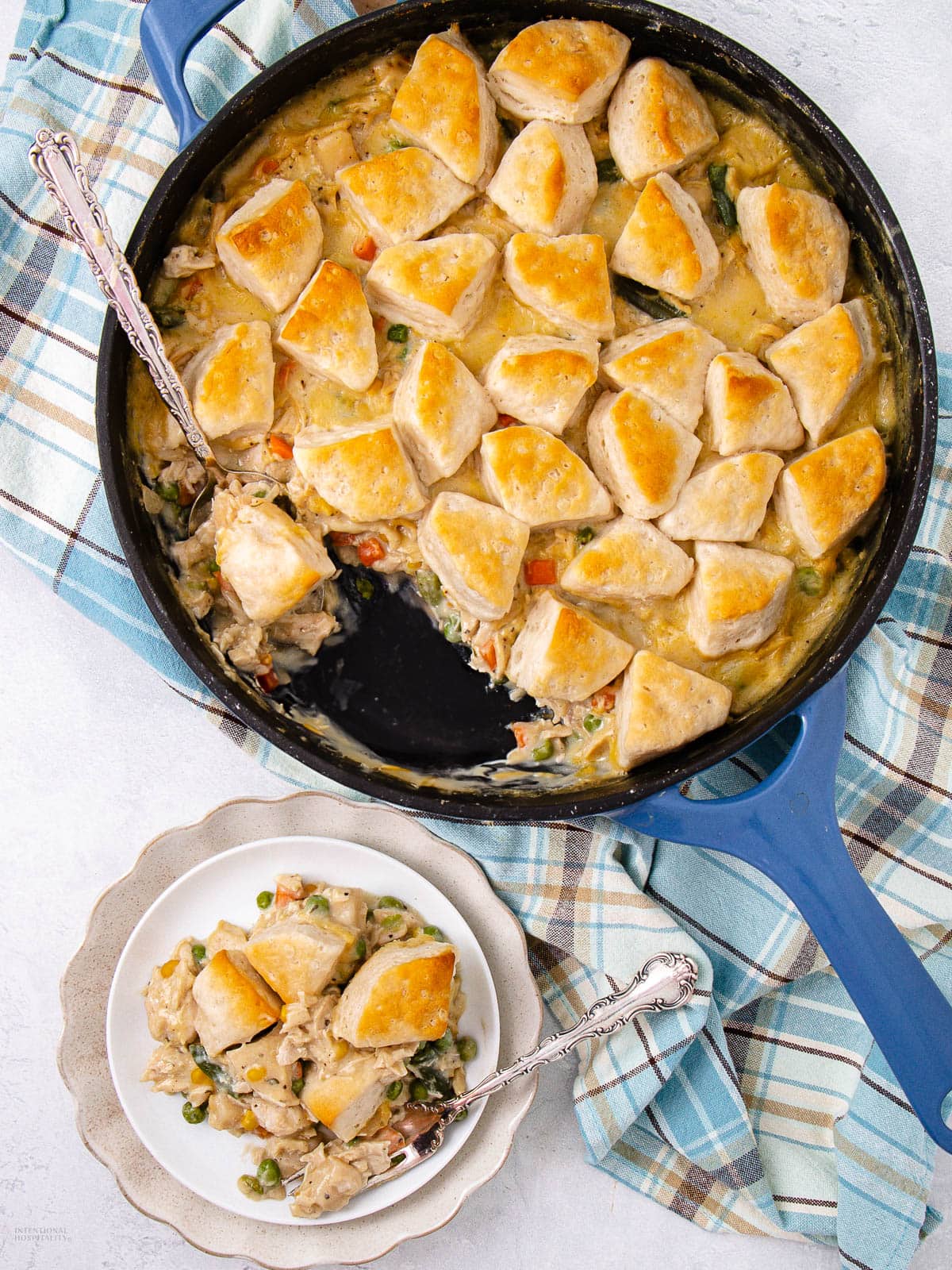 A skillet filled with creamy chicken and vegetable pot pie, topped with golden biscuit pieces. A serving has been scooped out onto a plate, with a spoon beside it. The skillet rests on a blue plaid towel.
