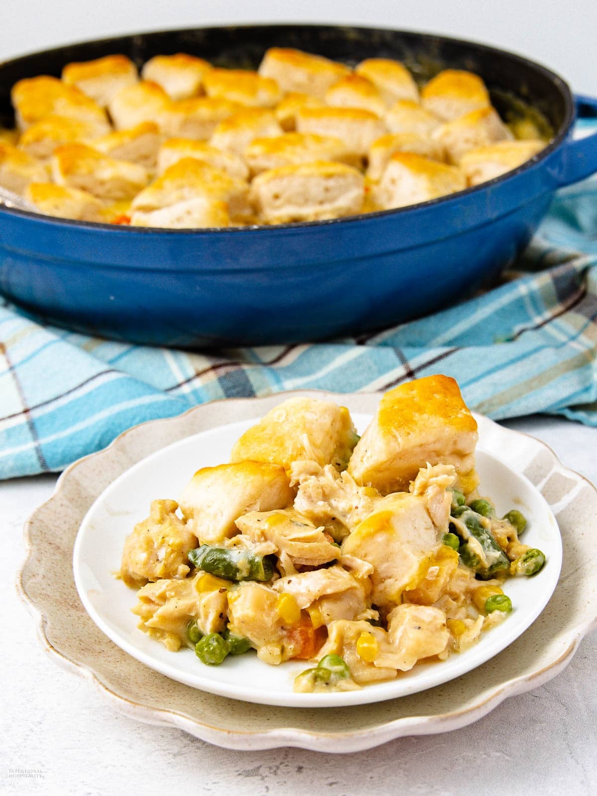 A plate of creamy chicken and vegetable casserole topped with golden biscuit pieces, served in front of a casserole dish with more of the same dish. The dish sits on a blue plaid cloth.