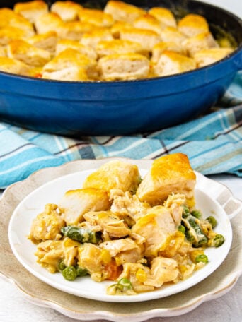 A white plate holds a serving of creamy chicken and vegetable casserole topped with golden biscuit pieces. In the background, the rest of the casserole sits in a blue baking dish on a plaid cloth.
