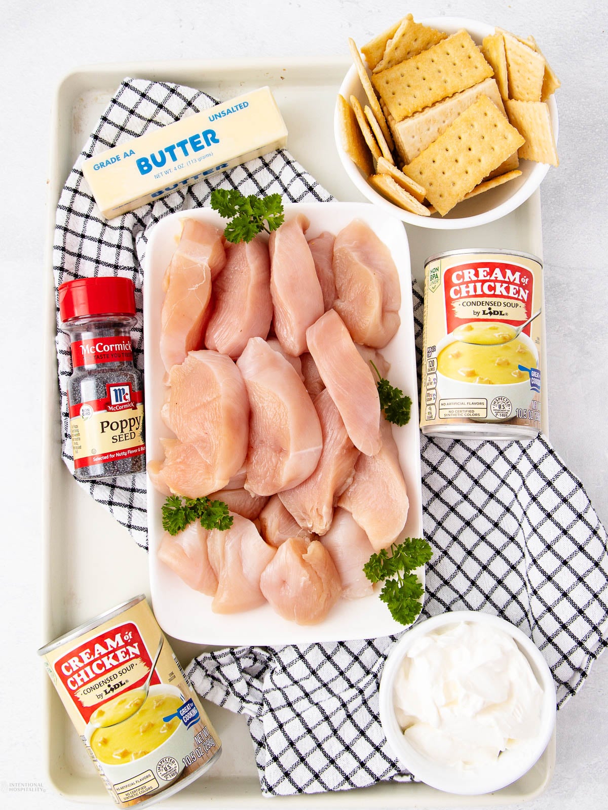 A tray with raw chicken breasts, two cans of cream of chicken soup, a bowl of sour cream, a bowl of crackers, a stick of butter, a jar of poppy seeds, and sprigs of parsley on a black-and-white towel.