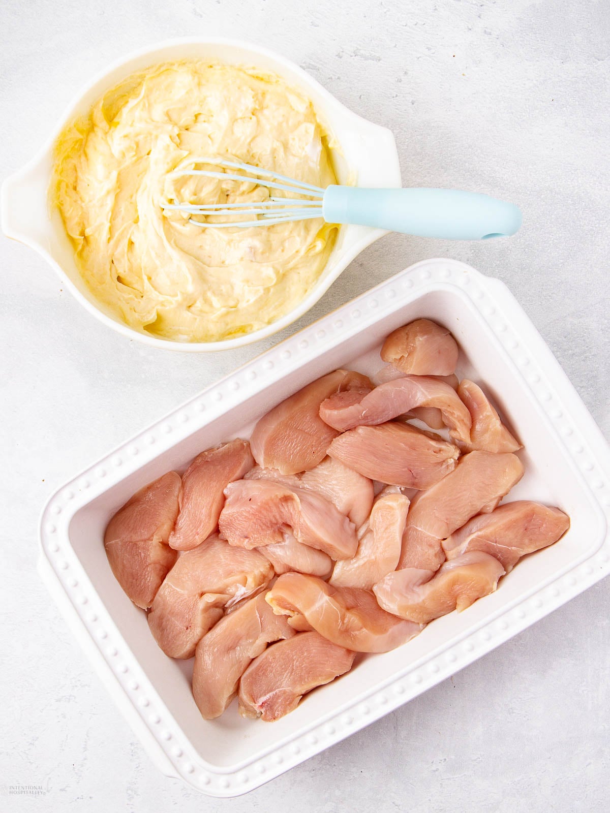 A white dish with raw chicken strips next to a bowl of creamy yellow mixture being whisked, on a light surface.