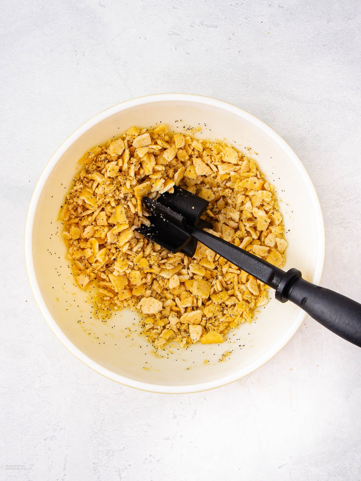 A white bowl filled with crushed crackers and seasonings is being mixed with a black spatula on a light-colored surface.