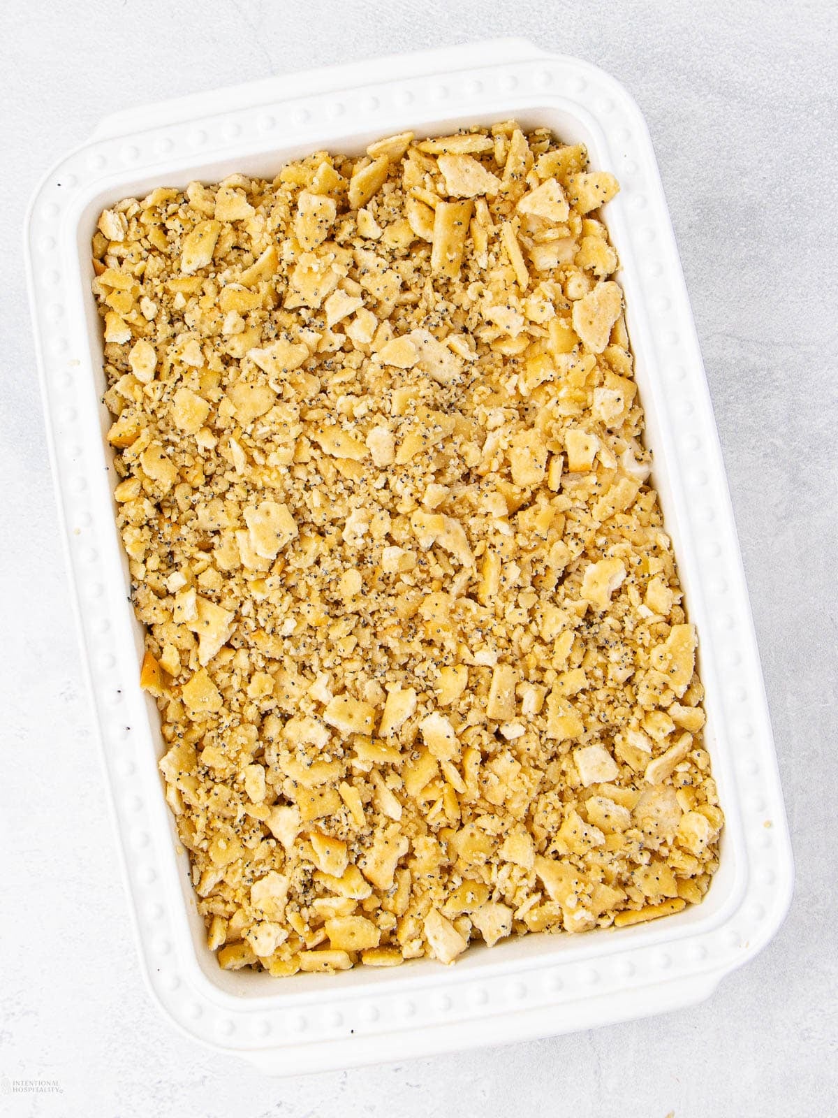 A white rectangular baking dish filled with a crumbly topping made of crushed crackers and spices, on a light gray surface.