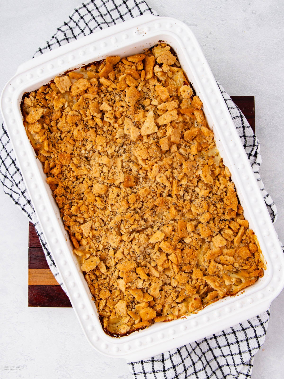 A white rectangular baking dish filled with a golden-brown, crumbly casserole topping, placed on a black and white checkered cloth and a wooden board.