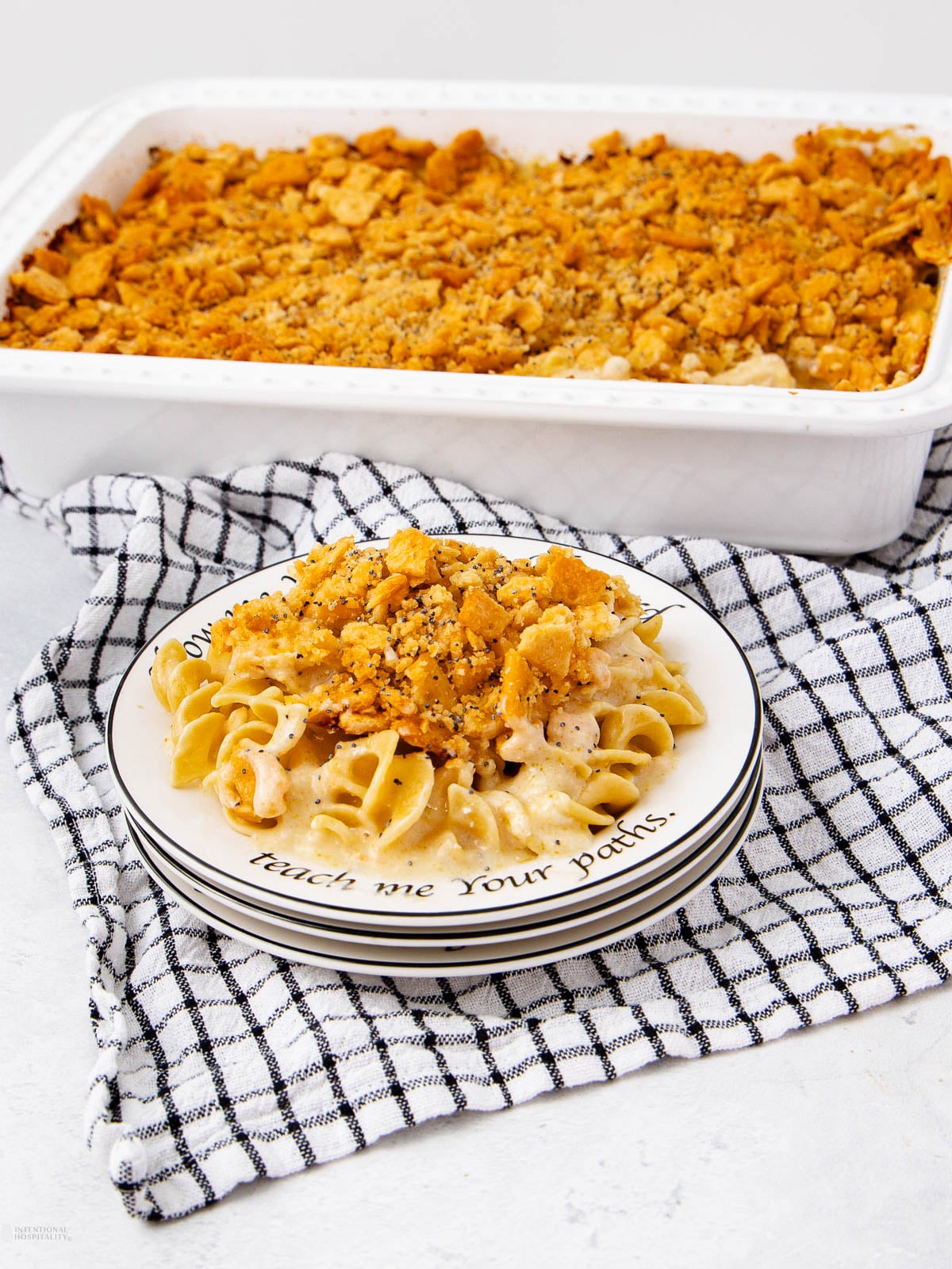 A plate of creamy noodle casserole topped with a golden, crunchy crumb topping sits on a black-and-white checkered towel, with a baking dish of the same casserole in the background.