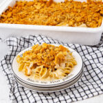 A plate of creamy noodle casserole topped with crushed crackers sits in front of a casserole dish on a black-and-white checkered towel. The plate has a decorative rim with text.