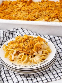 A plate of creamy noodle casserole topped with crushed crackers sits in front of a casserole dish on a black-and-white checkered towel. The plate has a decorative rim with text.
