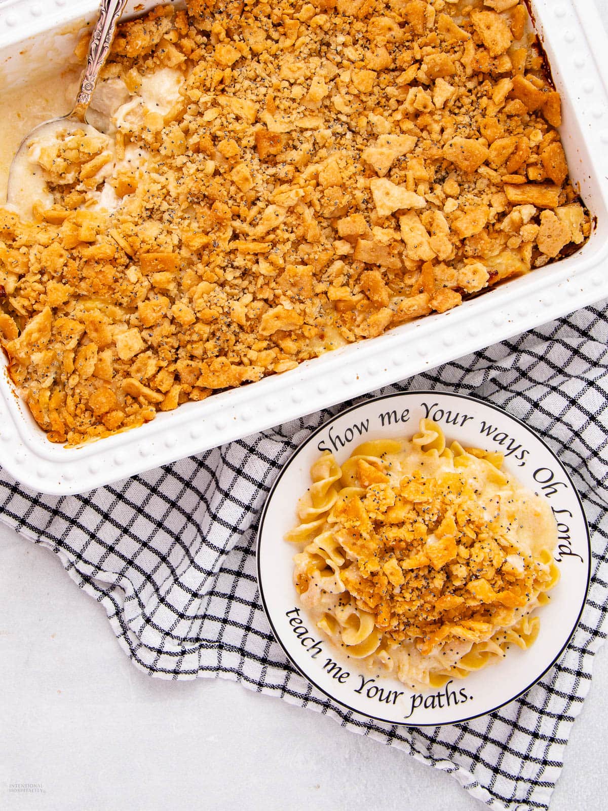A baking dish filled with a creamy casserole topped with crushed crackers sits on a checkered cloth. A serving is plated on a decorative plate with noodles and crumbled topping, beside the baking dish.