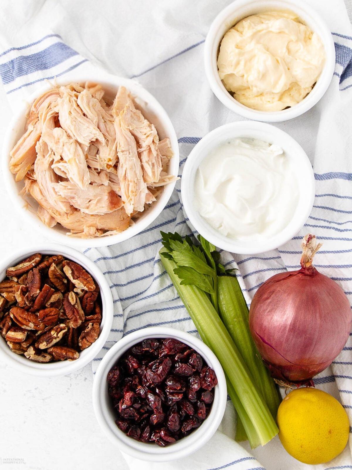 A top-down view of ingredients on a striped cloth: shredded chicken, mayonnaise, yogurt, chopped pecans, dried cranberries, celery stalks, a red onion, and a lemon.