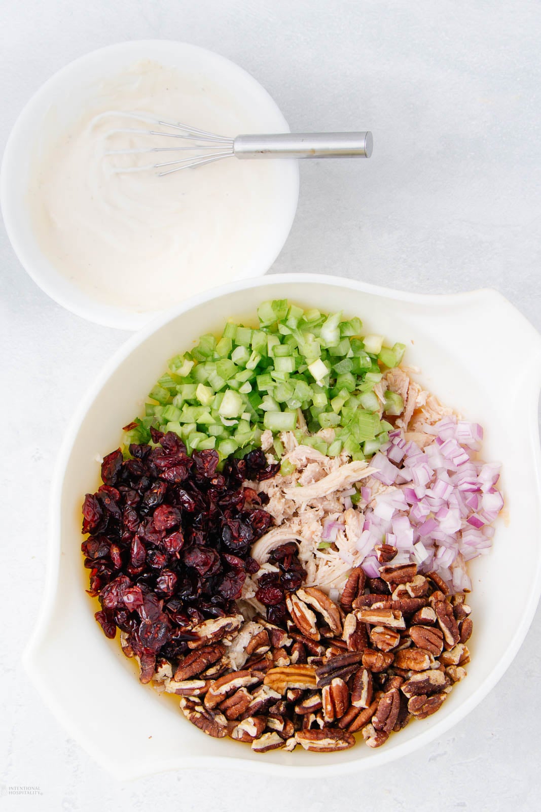 A large bowl contains chopped celery, diced red onion, shredded chicken, dried cranberries, and pecan pieces arranged separately. Nearby, a smaller bowl holds a creamy dressing with a whisk.