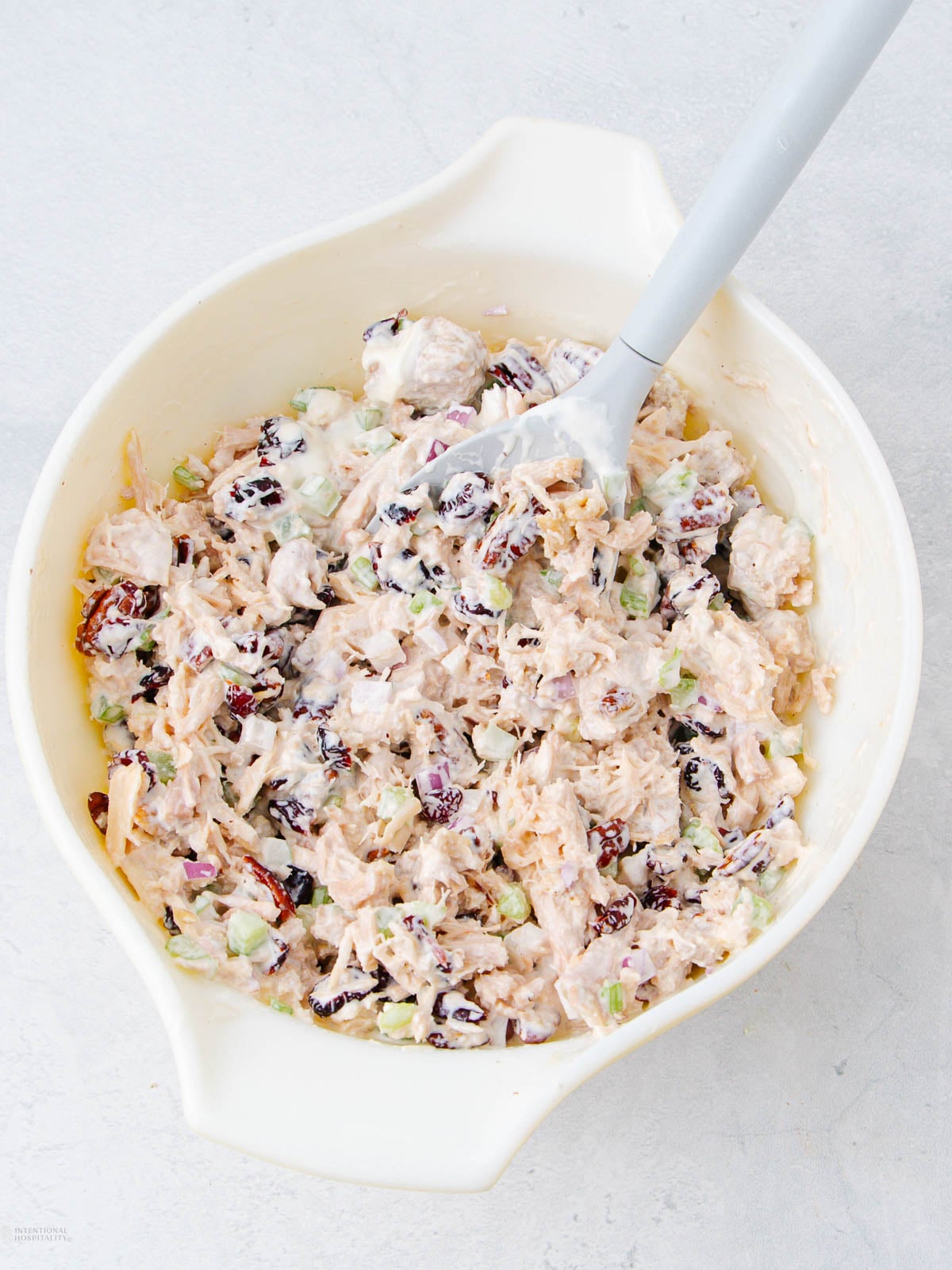 A white mixing bowl filled with chicken salad containing shredded chicken, celery, dried cranberries, and a creamy dressing, with a gray spatula resting inside.