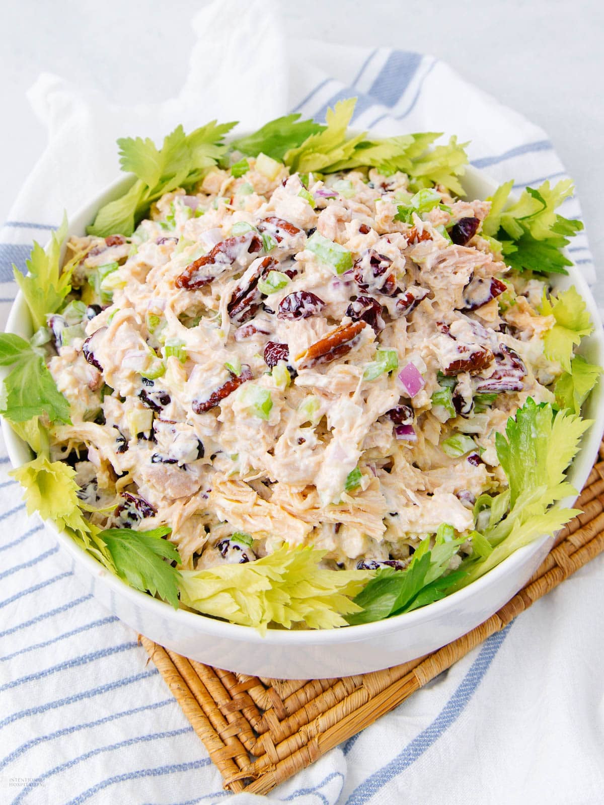 A bowl of chicken salad garnished with celery leaves, containing shredded chicken, pecans, dried cranberries, chopped celery, and red onion, on a striped towel and wicker mat.