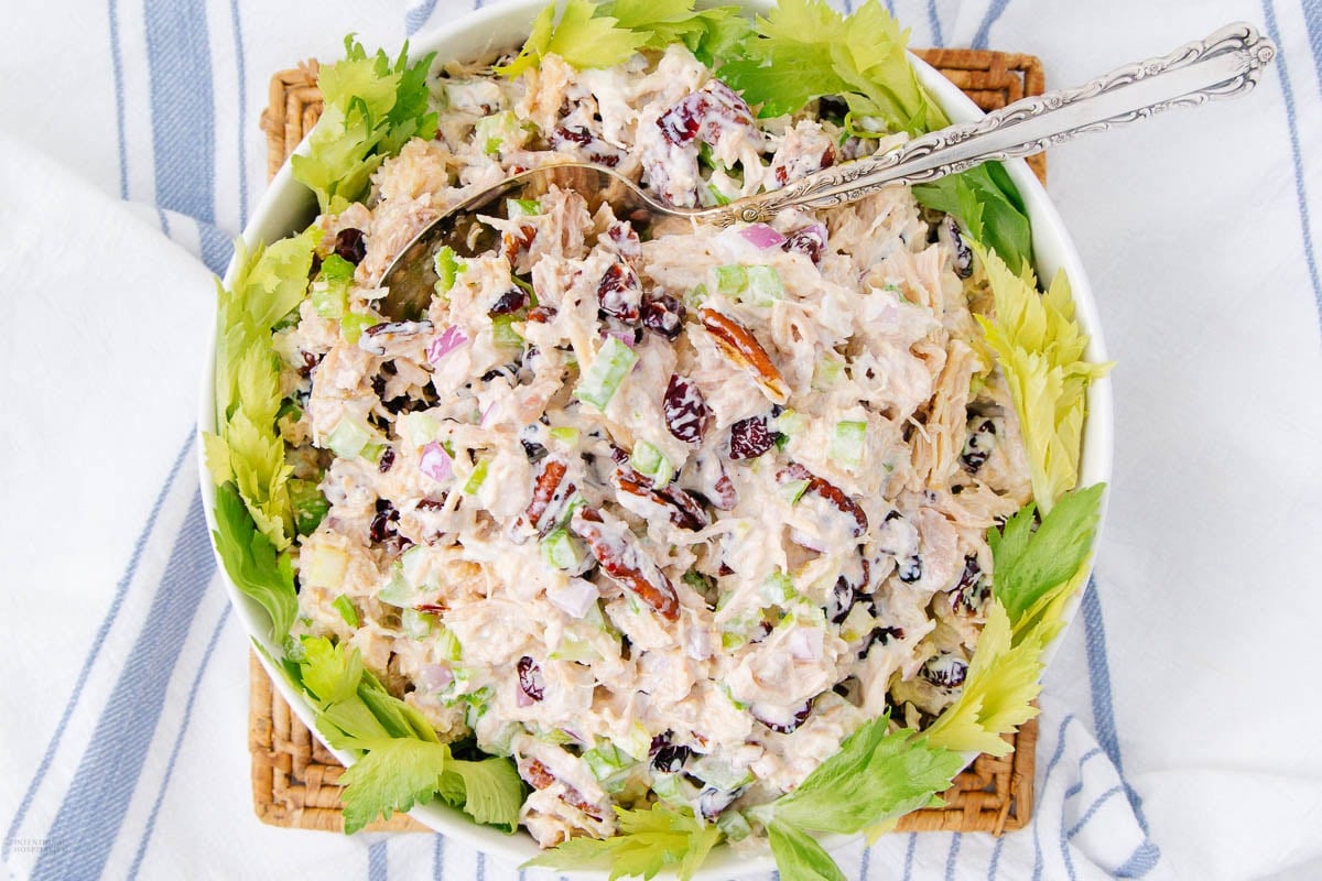 A bowl of chicken salad with celery, dried cranberries, and pecans, mixed with creamy dressing, garnished with celery leaves and served with a silver spoon on a woven placemat.