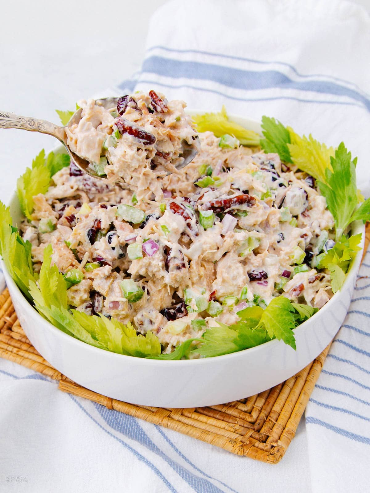 A bowl of creamy chicken salad mixed with celery, dried cranberries, and chopped nuts, garnished with celery leaves. A spoon holding a scoop of the salad is above the bowl, and the dish sits on a woven mat.