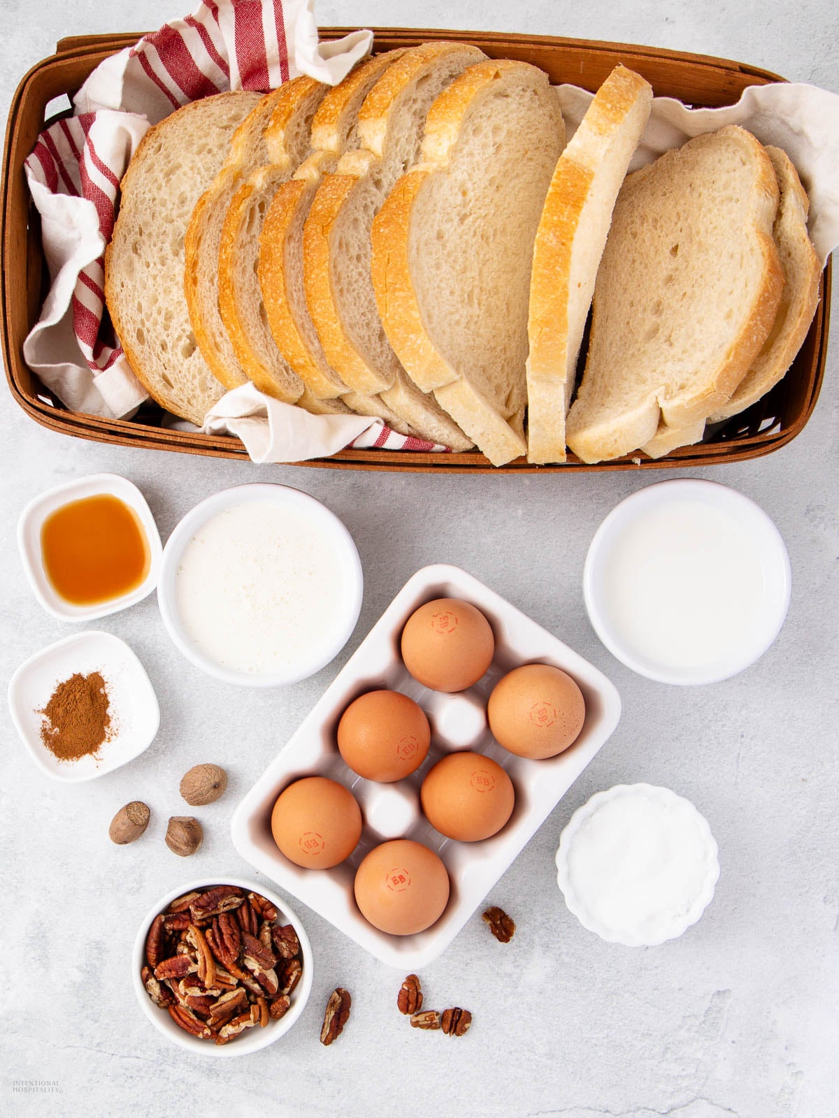 A basket of sliced bread, eggs in a carton, and small bowls with milk, cream, pecans, cinnamon, nutmeg, sugar, and vanilla extract, all arranged on a light surface for baking or cooking.