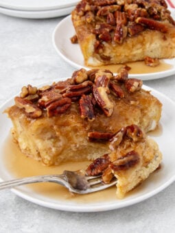 A slice of baked French toast topped with chopped pecans and syrup sits on a white plate with a fork. Another plate with a similar slice is visible in the background.