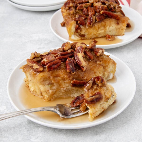 A slice of baked French toast topped with chopped pecans and syrup sits on a white plate with a fork. Another plate with a similar slice is visible in the background.