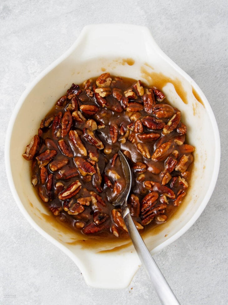 A white bowl filled with a mixture of pecans and thick brown syrup, with a metal spoon resting inside. The bowl is placed on a light gray surface.