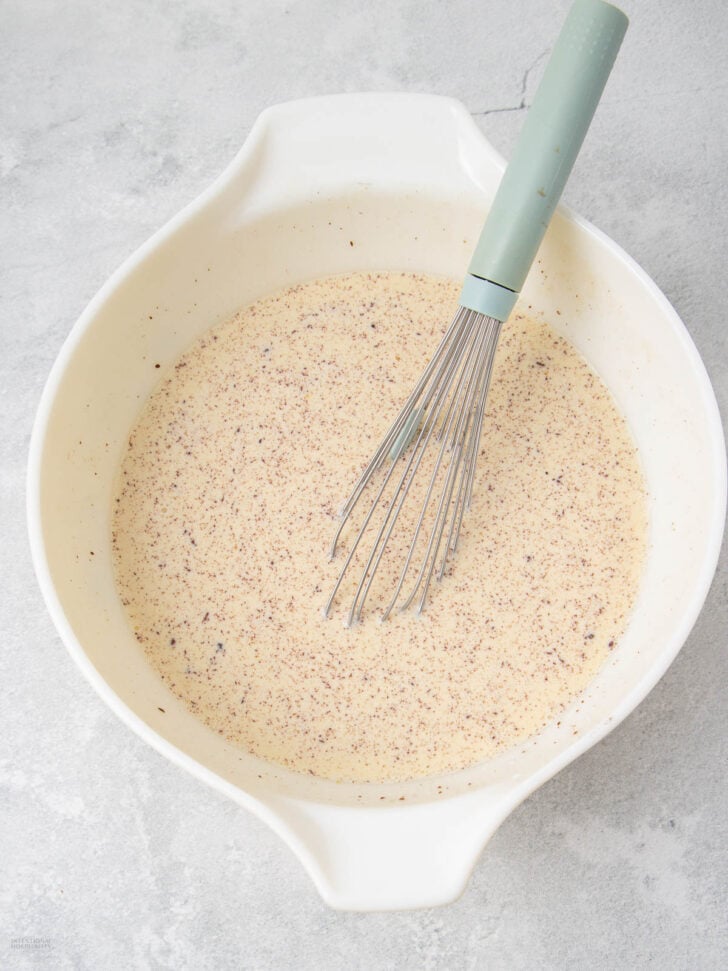 A white mixing bowl filled with a creamy, speckled liquid mixture, with a metal whisk featuring a light green handle resting inside the bowl on a light gray surface.