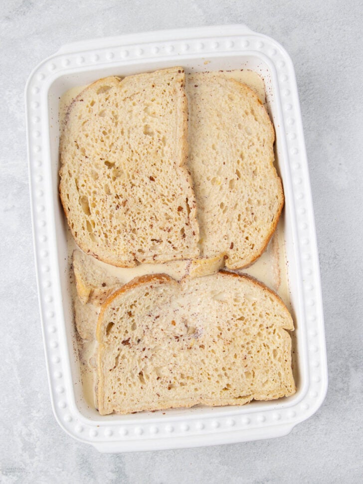 Four slices of bread are arranged in a white rectangular baking dish, partially soaked in a creamy liquid mixture, on a light gray surface.