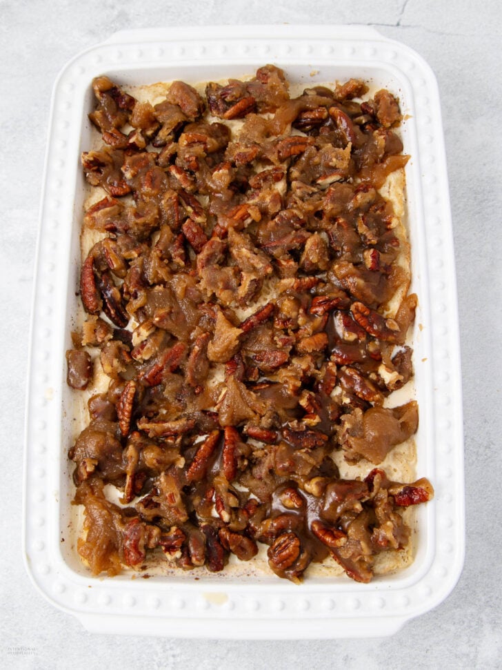 A white rectangular baking dish filled with a baked dessert topped with chopped pecans and a caramel-like glaze, resting on a light gray surface.