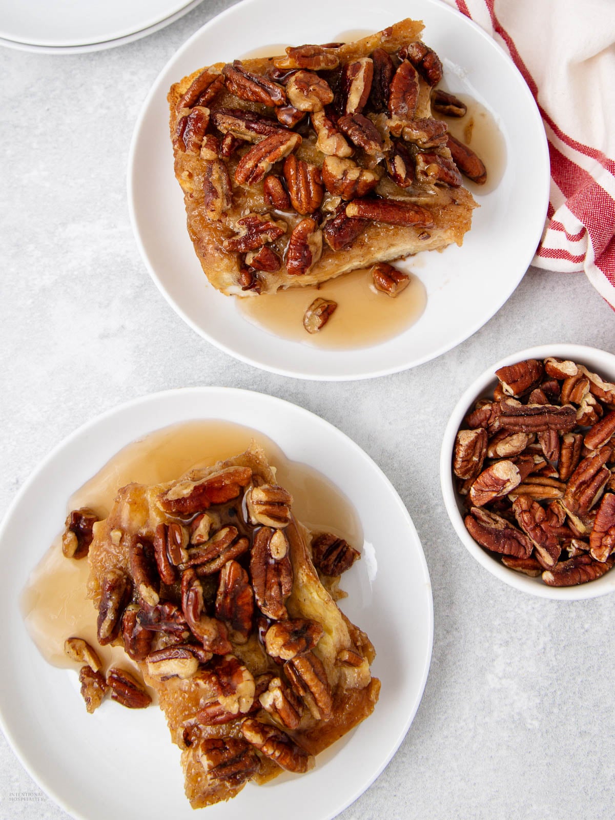 Two white plates each hold a serving of bread pudding topped with pecans and syrup. A small bowl filled with pecans sits nearby on a light-colored surface, and a red-and-white striped cloth is partially visible in the corner.