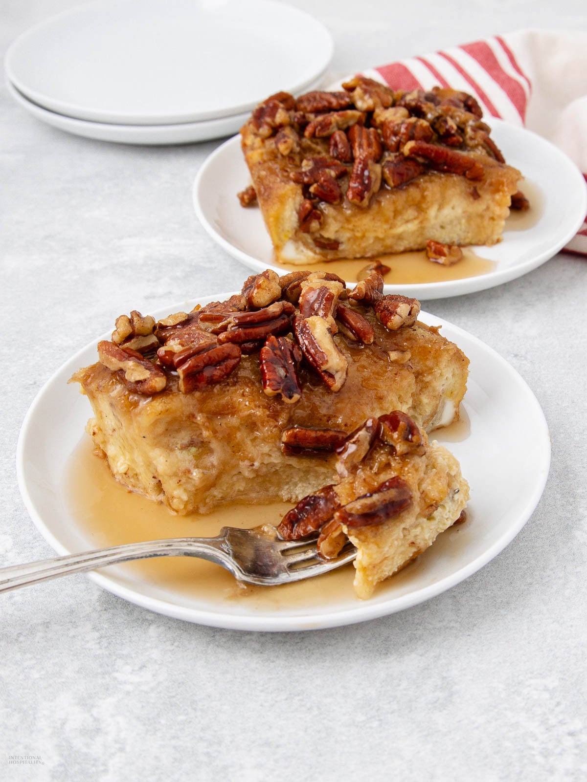 Two slices of bread pudding topped with chopped pecans and syrup on white plates, one with a fork. A red-striped napkin is in the background.