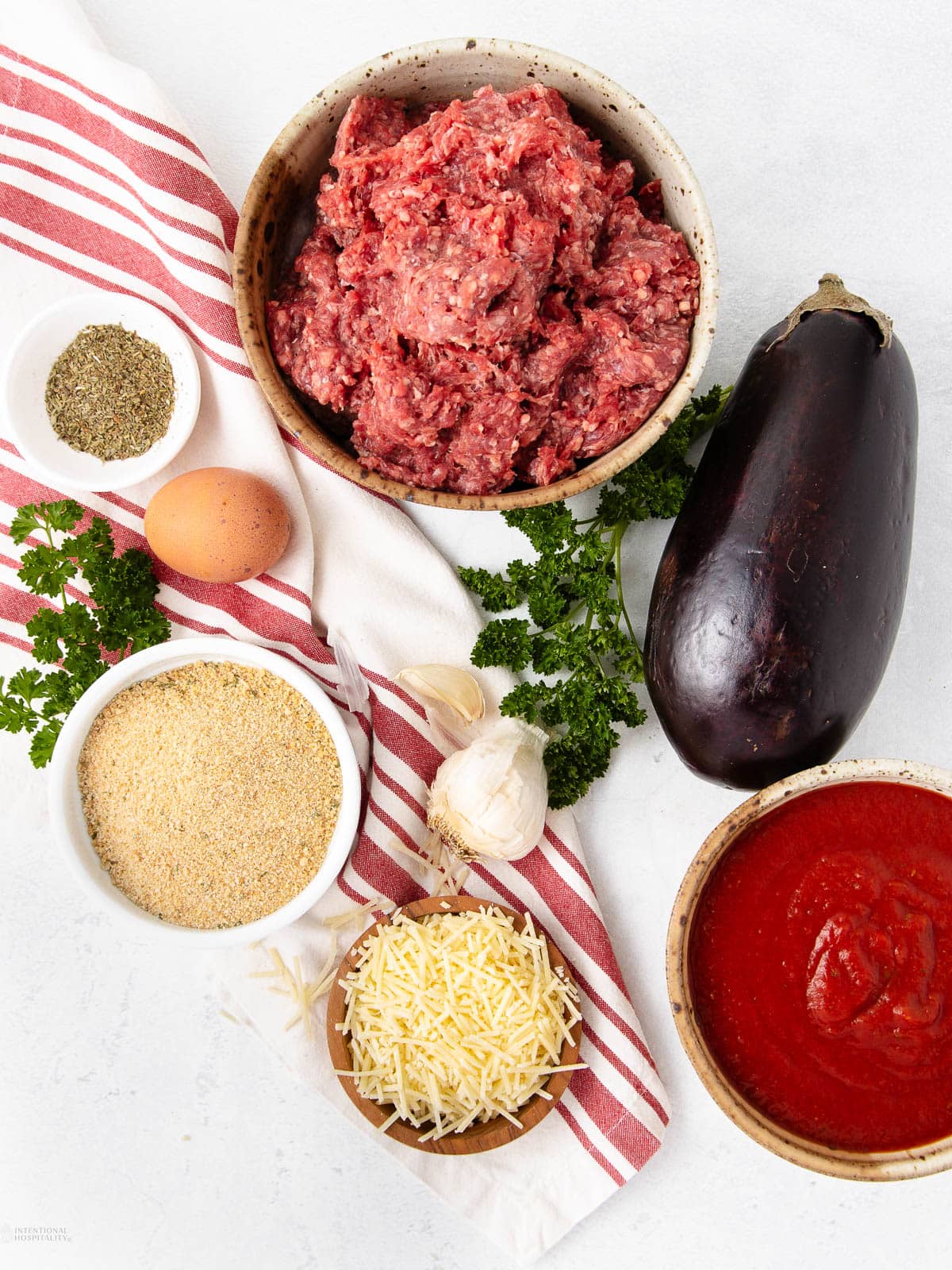 Flat lay of ingredients on a white surface: a bowl of ground meat, an eggplant, a bowl of marinara sauce, breadcrumbs, shredded cheese, an egg, garlic, parsley, dried herbs, and a red-striped kitchen towel.