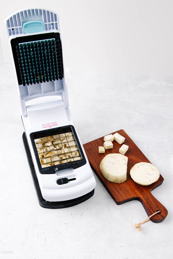 A vegetable chopper with diced pieces of root vegetable sits next to a wooden cutting board holding sliced and cubed pieces of the same vegetable on a light surface.