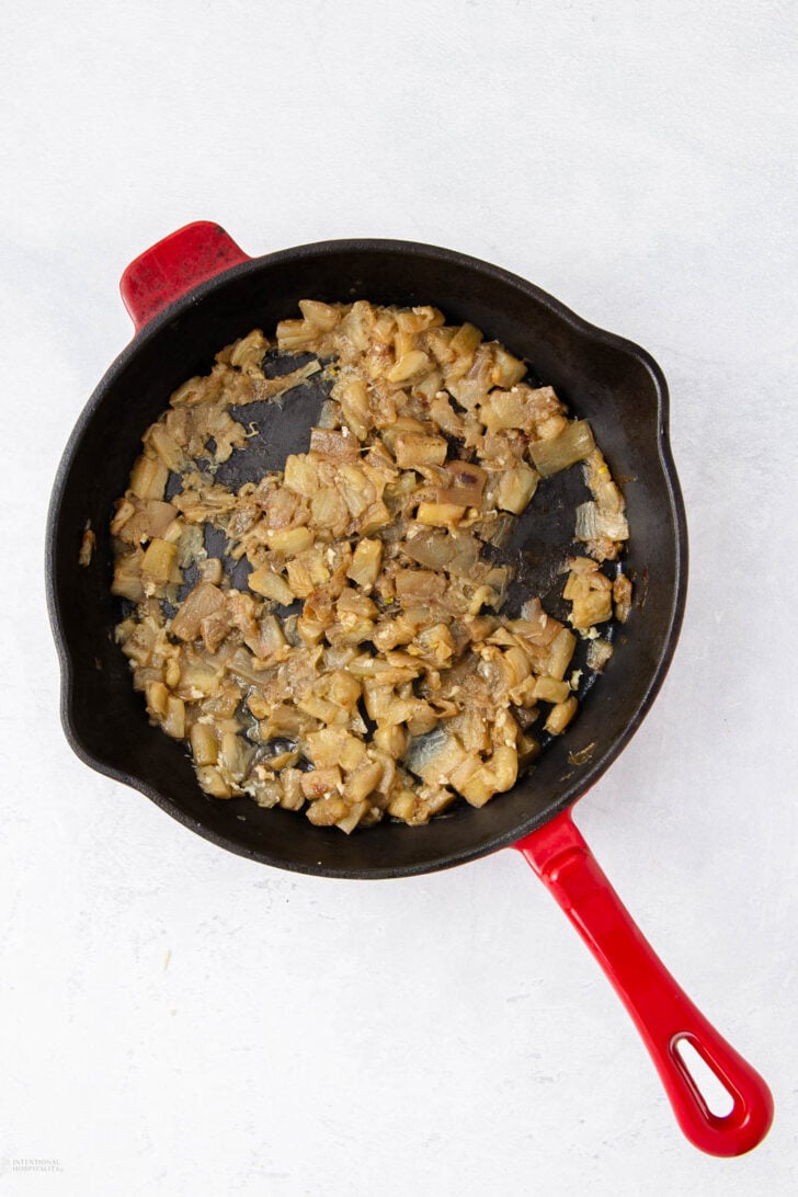 A black cast iron skillet with a red handle contains cooked, diced onions and eggplant on a light, textured surface.