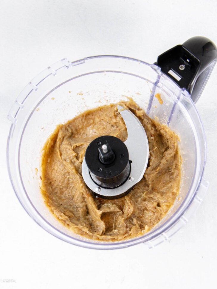 A food processor bowl containing a creamy, light brown mixture with a metal blade in the center, set on a white surface.