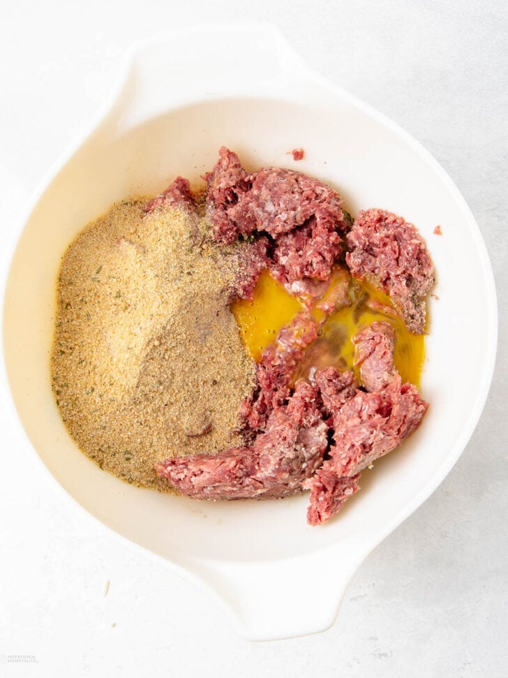A white mixing bowl containing raw ground meat, breadcrumbs, and a cracked egg on a light countertop.
